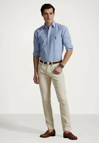 Man som bär en ljusblå knapp-up skjorta med fickemblem, beige byxor, brunt bälte, bruna loafers och armbandsur, står med handen i fickan.