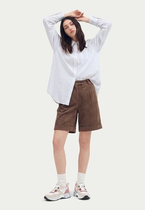 Femme portant une chemise ample rayée boutonnée, un short marron jusqu'aux genoux, des chaussettes blanches et des baskets rose et blanc, debout les bras levés au-dessus de la tête.