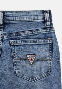 Niebieskie dżinsy o charakterystycznym kształcie kieszeni w formie trójkąta; czerwona i biała naszywka logo "GUESS" przyszyta do tylnej kieszeni.