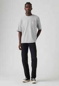 Muškarac stoji s pletenom frizurom, nosi svijetlosivu majicu, crne hlače i crno-bijele tenisice, nasuprot jednostavne pozadine.