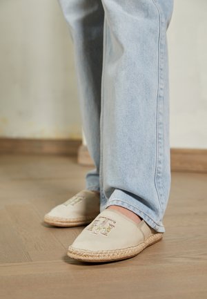 Fødder iført lyseblå jeans og beige slip-on espadrillos med flettede såler og dekorative initialer på trægulv.