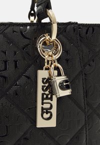 Sac à main noir en matériau synthétique texturé, présentant un motif en diamants, une quincaillerie dorée, un verrou et une étiquette avec le logo "GUESS".
