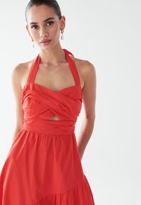 Punane kaela taha seotud kleit kokkupandud rinnakuga, lõhikuga detailide ja voolava seelikuga. Valmistatud kergest kangast, millel on sile tekstuur.