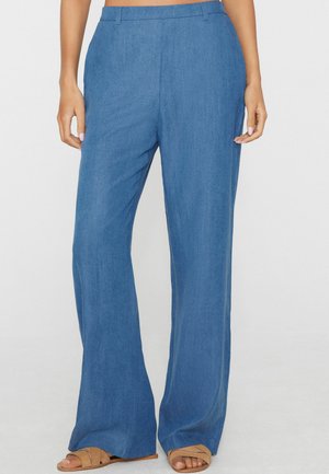 Frau trägt blaue Hose mit hoher Taille und weitem Bein sowie braune flache Sandalen, steht vor einem schlichten hellen Hintergrund.