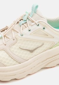 Sapatilha de corrida em bege claro com um upper de malha texturizada, detalhes em prateado, cordões elásticos e uma sola almofadada em verde pastel.