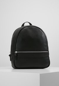 Svart läderryggsäck med en framficka med dragkedja, rundad topp och bärhandtag, placerad på en vit piedestal mot en grå bakgrund.