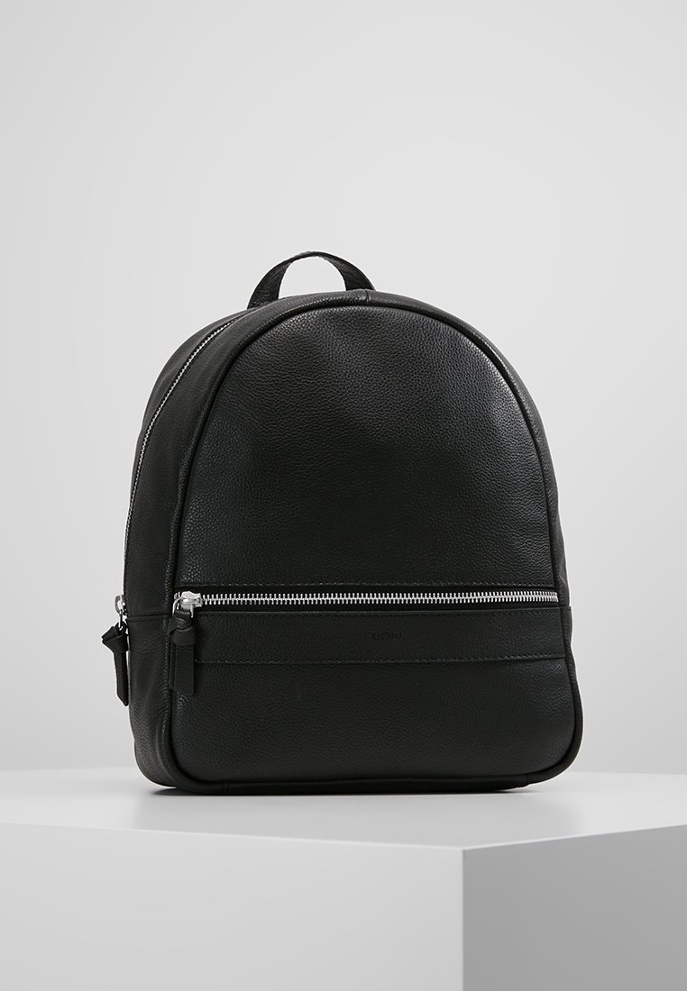 Svart läderryggsäck med en framficka med dragkedja, rundad topp och bärhandtag, placerad på en vit piedestal mot en grå bakgrund.