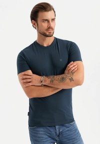 Námořnické modré tričko v přiléhavém designu s kulatým výstřihem, vyrobené z hladkého materiálu. Má malé logo na hrudi a krátké rukávy.