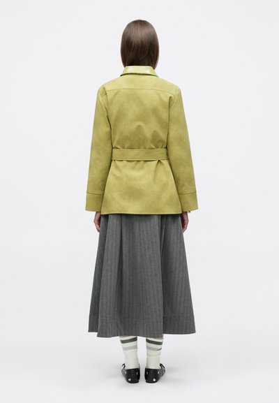 Γυναίκα με ίσια καστανά μαλλιά, φοράει ένα χακί μπουφάν με ζώνη, μακριά γκρι πλισέ φούστα, ριγέ κάλτσες και μαύρα παπούτσια, στέκεται με την πλάτη προς τα εμπρός.