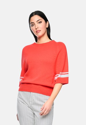 Frau trägt einen hellorangen Strickpullover mit weißen Streifen an den Ärmeln und weiße Hosen mit vertikalen schwarzen Streifen und steht vor einem schlichten Hintergrund.