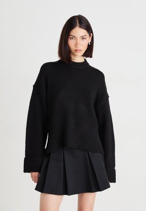 Young woman with straight dark hair wearing pearl earrings, a loose black sweater, and a pleated black skirt against a plain background.
