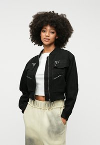Črna bomber jakna z kovinskimi dodatki, zadrgo spredaj in dvema žepoma na prsih; v kombinaciji s svetlo obarvanimi, ohlapno prilegajočimi se trenirkami.