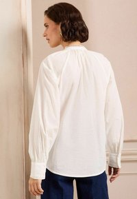Blusa bianca a maniche lunghe con design plissettato al colletto, polsini con bottoni e una vestibilità rilassata, abbinata a pantaloni blu scuro.