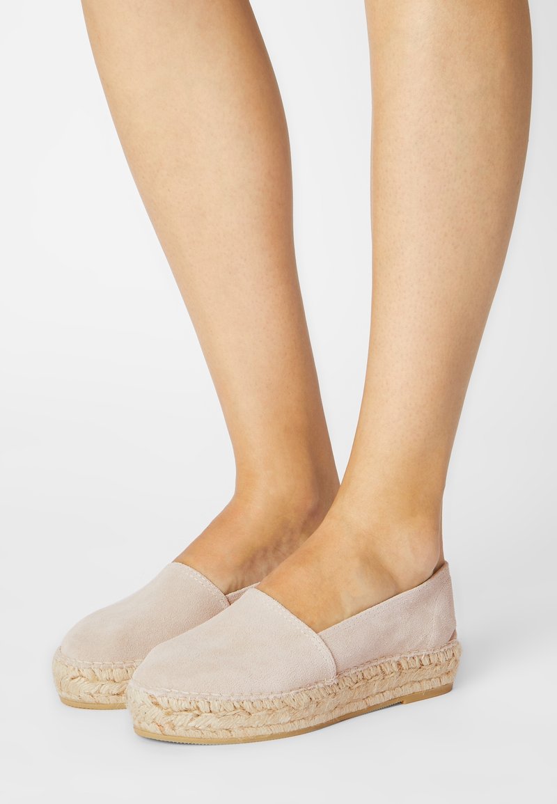 Pieds portant des espadrilles à enfiler en suède beige clair avec des semelles en jute tressé sur un fond blanc.