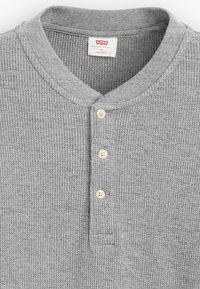 Camiseta henley gris con relieve y cuello redondo, que presenta tres botones de madera en la tapeta y una etiqueta en la zona del cuello.