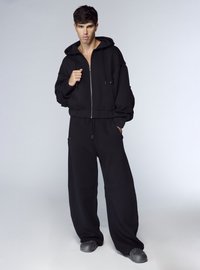 Felpa nera con zip frontale, vestibilità ampia e maniche oversize abbinata a pantaloni neri a gamba larga coordinati. Tessuto strutturato, dettagli minimi.