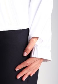 Ręka z naturalnymi paznokciami spoczywa na czarnych spodniach, ubrana w białą koszulę z długim rękawem zakończoną elastycznym mankietem na nadgarstku.