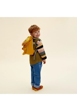 Junger Junge mit roten Haaren, der einen gestreiften Pullover, eine grüne Weste, blaue Jeans und orangefarbene Schuhe trägt und einen gelben Rucksack in Löwenform bei sich hat.