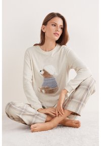Top beige claro de manga larga con un diseño de oso de peluche que lleva un suéter azul y un gorro blanco. La modelo viste pantalones de pijama a cuadros en tonos suaves.