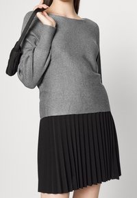 Maglione grigio a coste con scollatura ampia abbinato a una gonna plissettata nera. L'outfit presenta una parte superiore texturizzata e una parte inferiore liscia.