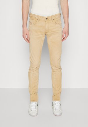 Beige Slim-Fit-Chinos mit einer subtilen Textur, fünf Taschen und metallenen Beschlägen. Kombiniert mit weißen Sneakers mit schwarzen Akzenten.