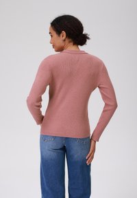 Ribotė, pritaikyta, ilgomis rankovėmis palaidinė dulkiai rožinės spalvos su apvalia iškirpte, derinama su aukštu liemeniu mėlynomis plačiomis kelnių, matoma iš nugaros.