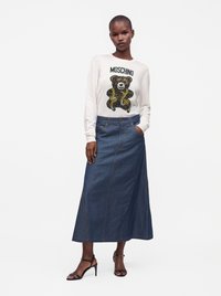 Femme portant un pull blanc Moschino avec un motif ourson et une longue jupe en jean bleue, debout avec une main dans la poche et des talons noirs.