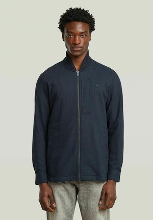 Navy zip-up jacket with a ribbed collar, two front pockets, textured fabric, and a rectangular patch on the chest for branding.