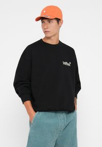 Zwarte crewneck sweatshirt met een losse pasvorm, voorzien van een klein wit logo. Gecombineerd met lichtblauwe, gestructureerde shorts en een oranje pet.
