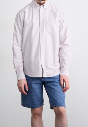 Homme portant une chemise rose clair boutonnée à manches longues et un short en denim bleu sur un fond uni.