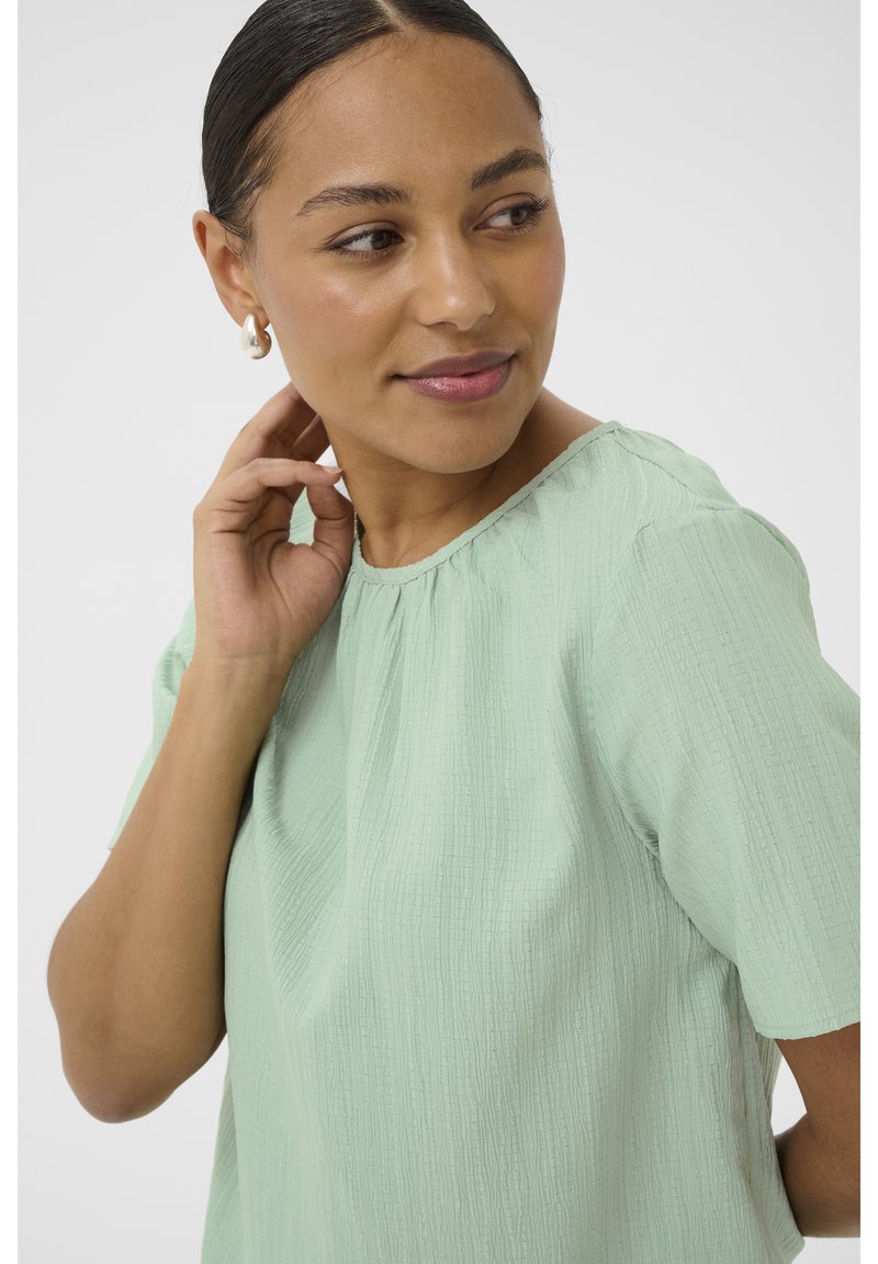 Femme aux cheveux foncés tirés en arrière portant des boucles d'oreilles en perles en forme de créoles et un chemisier texturé vert menthe à manches courtes, touchant son cou et regardant sur le côté.