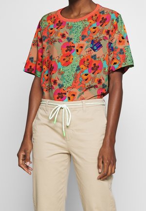 Personne portant une chemise à manches courtes fleurie et lumineuse avec des tons orange, rose, vert et bleu, associée à un pantalon beige et une ceinture blanche à cordon.
