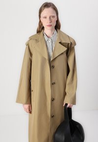 Beige trenchcoat med store reverser og seks knapper, båret over en svart og hvitstripet skjorte. Modellen holder en svart veske med en buet form.