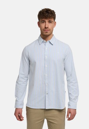 Mężczyzna ubrany w jasnoniebieską i białą koszulę z pionowymi paskami oraz spodnie w kolorze beżowym, stojący na tle jednolitego białego tła.