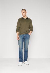 Oljčno zelena zip-up pulover, mehak material, dolge rokave, v kombinaciji z svetlo modrimi kavbojkami in belimi supergami. Preprost dizajn in prileganje.