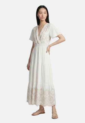 Femme portant une robe blanche jusqu'aux chevilles avec des manches courtes et des motifs floraux ajourés près de l'ourlet, associée à des sandales plates beiges.