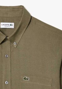 Camicia verde oliva con bottoni, realizzata in un tessuto testurizzato, vestibilità regolare e un piccolo logo di coccodrillo ricamato sul petto. Due chiusure a bottone sul colletto.