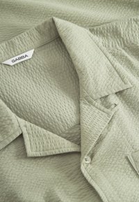 Svetlo zelena majica z teksturirano tkanino, ki ima ovratnik z ostrim robom, zapenjanje na gumbe in etiketo "GABBA" znotraj izreza za vrat.