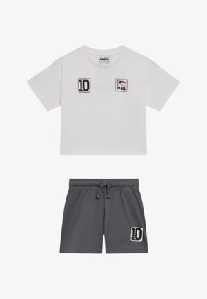 T-shirt bianca a maniche corte e corta con toppe nere e rosse, abbinata a pantaloncini grigio scuro con elastico in vita e toppa nera sulla gamba sinistra.