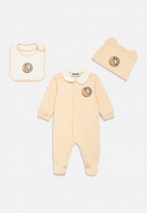 Beige Baby-Strampler mit weißem Kragen, passendem beigen Mützchen mit Bärenohren und weißem Lätzchen, jeweils mit Teddybär-Logo und Moschino-Branding.