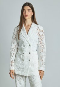Blazer bianco in pizzo con design doppiopetto, motivi floreali e bottoni argentati. Presenta maniche lunghe e semitrasparenti e una finitura textured.