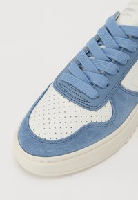 Blå fløyelsko og hvit perforert lær sneaker med blå snørebånd og kontrasterende beige gummisåle. Kjennetegnes av rund tå og sydd detaljer.