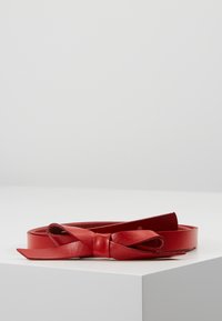 Cinto in pelle rossa con una texture liscia e un dettaglio a fiocco nella chiusura. Forma piatta ed allungata, che mostra un design minimalista.