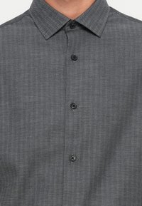 Camisa de botones gris con un patrón de espiga, que presenta un cuello puntiagudo y botones oscuros sobre una tela suave.