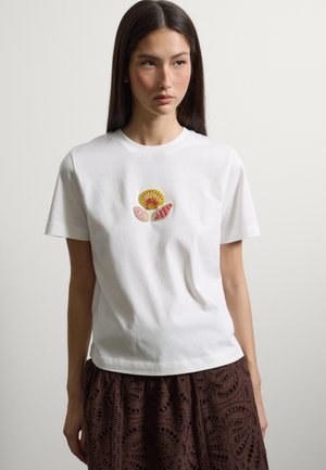 Junge Frau mit langen dunklen Haaren, die ein weißes T-Shirt mit drei bestickten Muscheln und einen dunkelbraunen gemusterten Rock trägt.