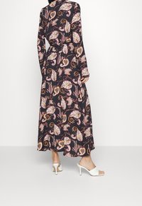 Žena v dlouhých černých šatech s barevným paisley potiskem a bílých sandálech s otevřenou špičkou na vysokém podpatku, postávající na neutrálním světlém pozadí.