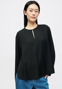Μαύρη μπλούζα σε χαλαρή γραμμή με στρογγυλή λαιμόκοψη, διαθέτει μπροστινή σχισμή και χρυσή λεπτομέρεια. Φτιαγμένη από λεία ύφασμα, με μακριά μανίκια και χαλαρή σχεδίαση.