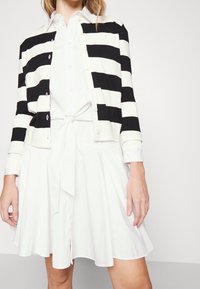 Czarny i biały pasiasty kardigan nałożony na białą sukienkę, z wiązaniem w talii, guzikowym zapięciem z przodu oraz długimi rękawami z podwiniętymi mankietami.