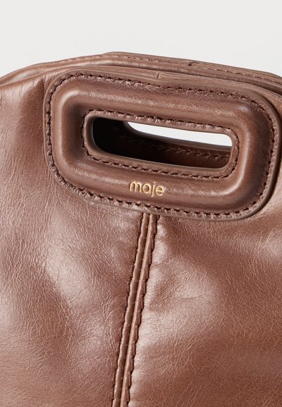 Κοντινή λήψη καφέ δερμάτινης τσάντας με ορθογώνια χειρολαβή και ανάγλυφο χρυσό λογότυπο "maje" κάτω από τη χειρολαβή.