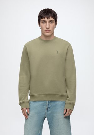 Junger Mann trägt einen einfarbigen olivgrünen Pullover und hellblaue Jeans, steht vor einem weißen Hintergrund.